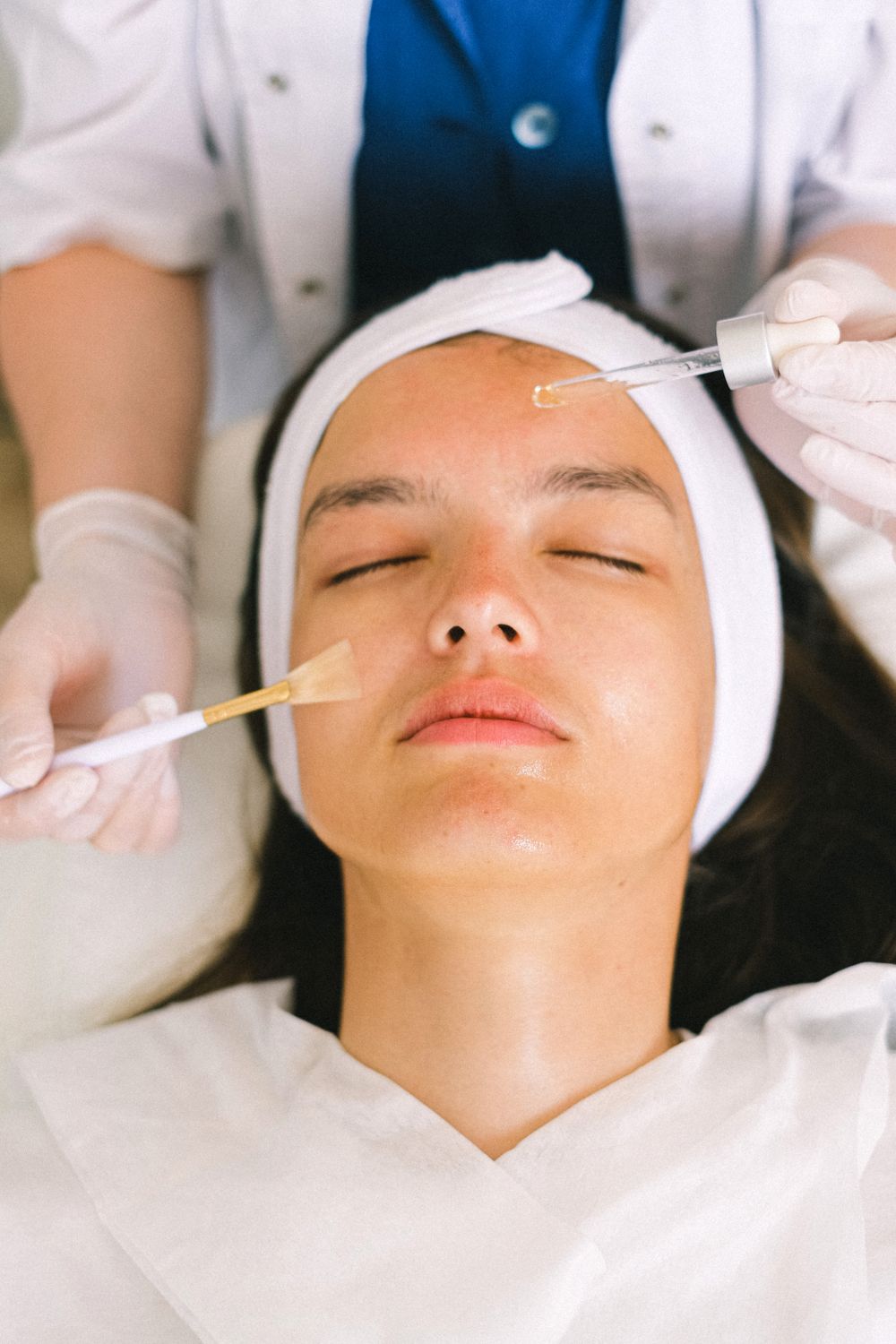 A client rests with eyes closed while a gloved provider applies serum during a facial treatment
